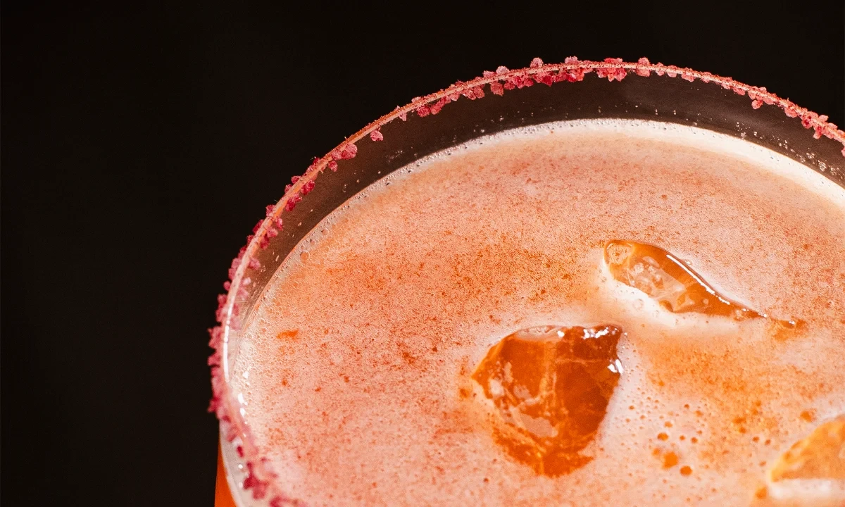 Drink autoral alaranjado em taça com borda de açúcar rosado e gelo translúcido