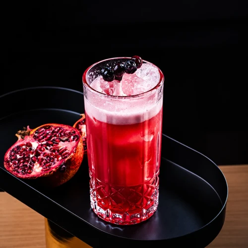 Grand Romã vermelho em copo alto de cristal, com espuma e frutas escuras, ao lado de uma romã aberta