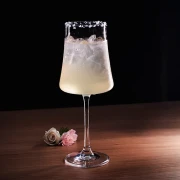 Taça alta de haste fina com drink claro, gelo e borda de açúcar, sobre mesa de madeira ao lado de duas rosas
