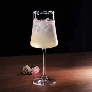 Taça alta de haste fina com drink claro, gelo e borda de açúcar, sobre mesa de madeira ao lado de duas rosas