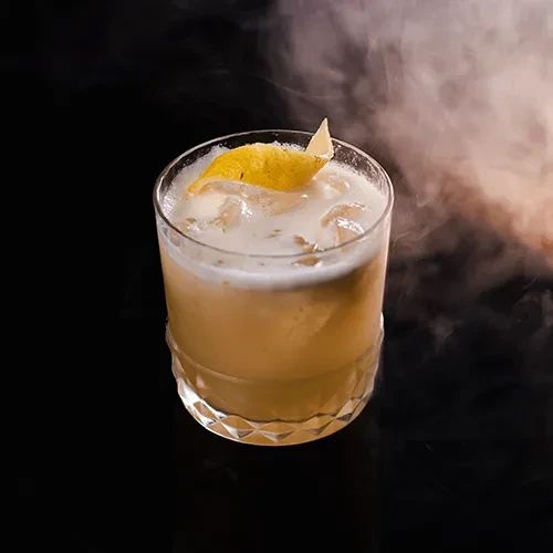 Copo baixo lapidado com drink amarelado, espuma, gelo e casca de limão, com fumaça sobre fundo escuro