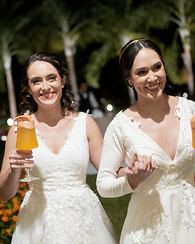 Duas mulheres de vestido branco sorrindo de mãos dadas, cada uma com uma taça de drink alaranjado, em um jardim à noite