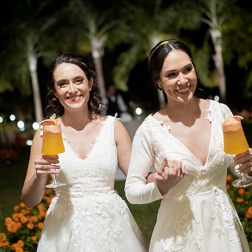 Duas mulheres de vestido branco sorrindo de mãos dadas, cada uma com uma taça de drink alaranjado, em um jardim à noite
