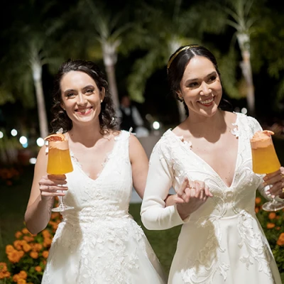 Duas mulheres de vestido branco sorrindo de mãos dadas, cada uma com uma taça de drink alaranjado, em um jardim à noite