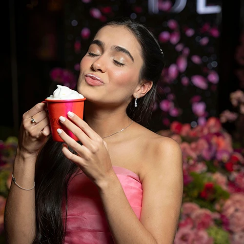 Jovem de vestido rosa, de olhos fechados, segurando um copo vermelho com espuma branca diante de uma parede de flores