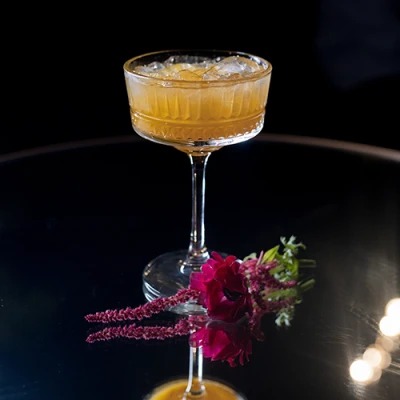 Taça coupé de cristal com drink âmbar e gelo sobre mesa espelhada, ao lado de uma flor vermelha e ramos de amaranto