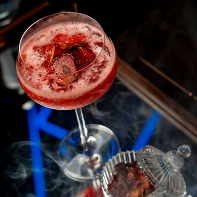 Taça coupé com drink vermelho espumoso e fatias de fruta desidratada, envolta em fumaça, com pote de cristal ao lado