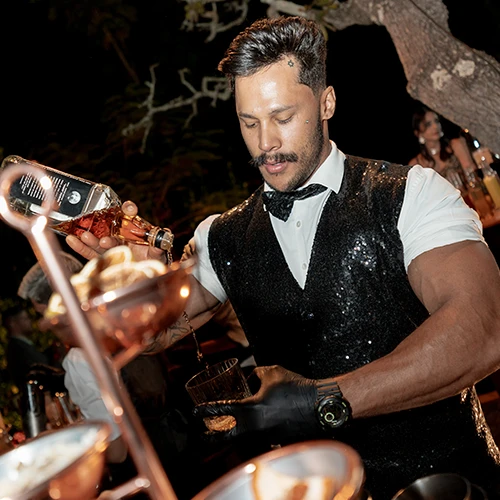 Bartender de colete de paetês e luvas pretas servindo uísque em um copo, à noite, ao ar livre