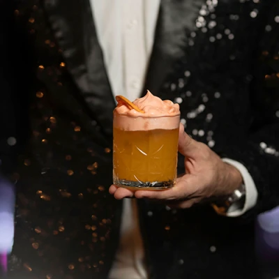 Mão segurando copo baixo lapidado com drink âmbar, espuma rosada e casca de laranja desidratada, diante de um paletó de paetês