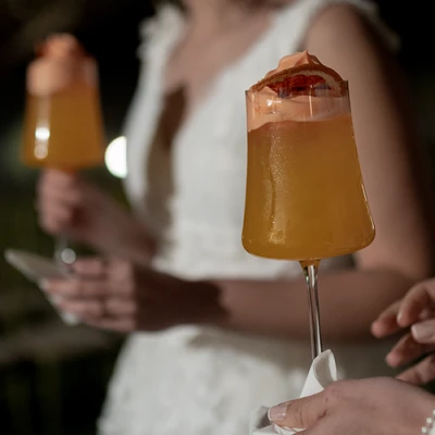 Mãos segurando taças altas com drink alaranjado, espuma e rodela de laranja desidratada, com uma mulher de vestido branco ao fundo