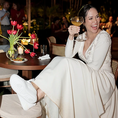 Mulher de vestido branco rindo e erguendo uma taça de drink dourado, sentada à mesa com tulipas vermelhas