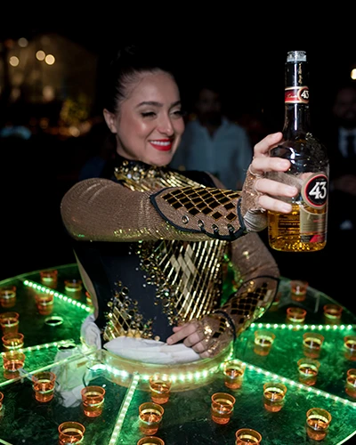 Mulher de figurino dourado vestindo uma mesa redonda iluminada, cheia de doses em copinhos, e segurando uma garrafa de licor