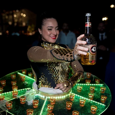 Mulher de figurino dourado vestindo uma mesa redonda iluminada, cheia de doses em copinhos, e segurando uma garrafa de licor
