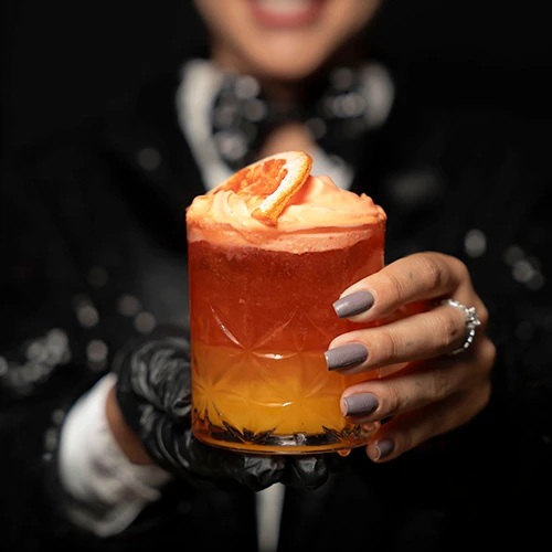 Mão estendendo um copo baixo lapidado com drink vermelho e laranja, espuma e rodela de laranja desidratada