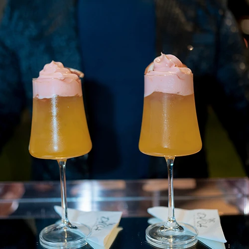 Duas taças altas com drink alaranjado e espuma rosa no topo, sobre guardanapos no balcão