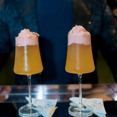 Duas taças altas com drink alaranjado e espuma rosa no topo, sobre guardanapos no balcão