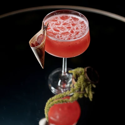 Taça coupé com drink vermelho espumoso e um cone de papel com fruta desidratada preso na borda, sobre mesa espelhada com amaranto verde
